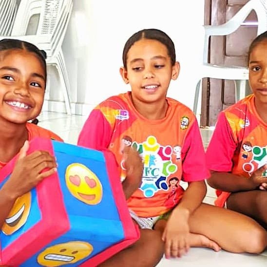 Três crianças sorridentes do projeto FFC sentadas no chão, uma segurando um cubo de papel com emojis.