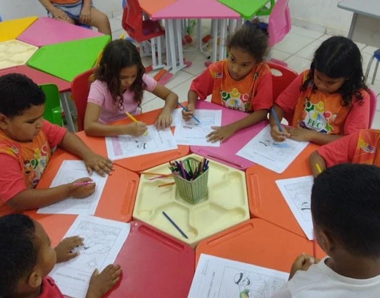 Crianças do projeto FFC fazem atividades de pintura ao redor de uma mesa hexagonal.