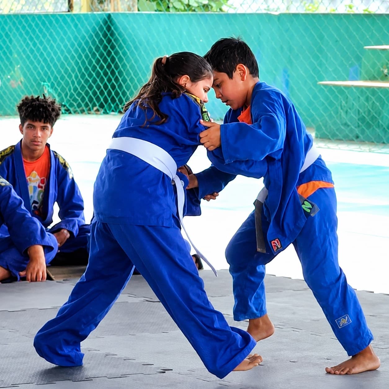 Duas crianças de quimono azul praticam judô sobre o tatame, com colegas observando ao fundo.