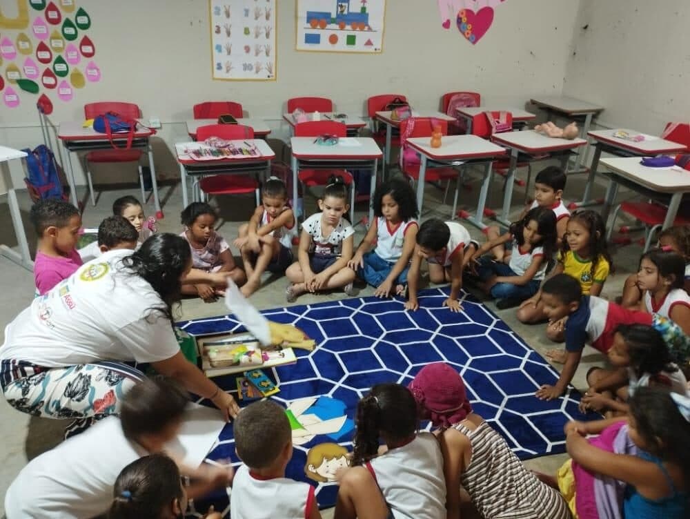 Educadora do FFC conduz uma roda de crianças sentadas no chão de uma sala, apresentando materiais da atividade.