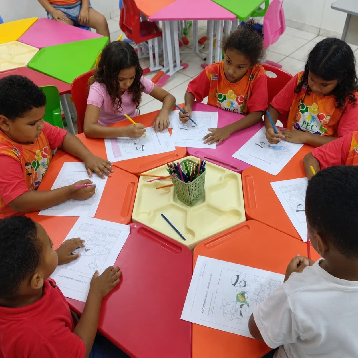 Crianças ao redor de mesas coloridas pintando desenhos, com camisetas laranja do projeto FFC.